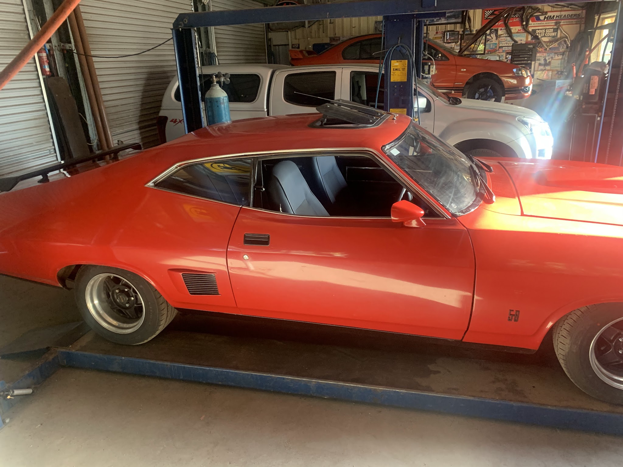 An orange XB Falcon coupe sitting on the hoist ramps inside the workshop
