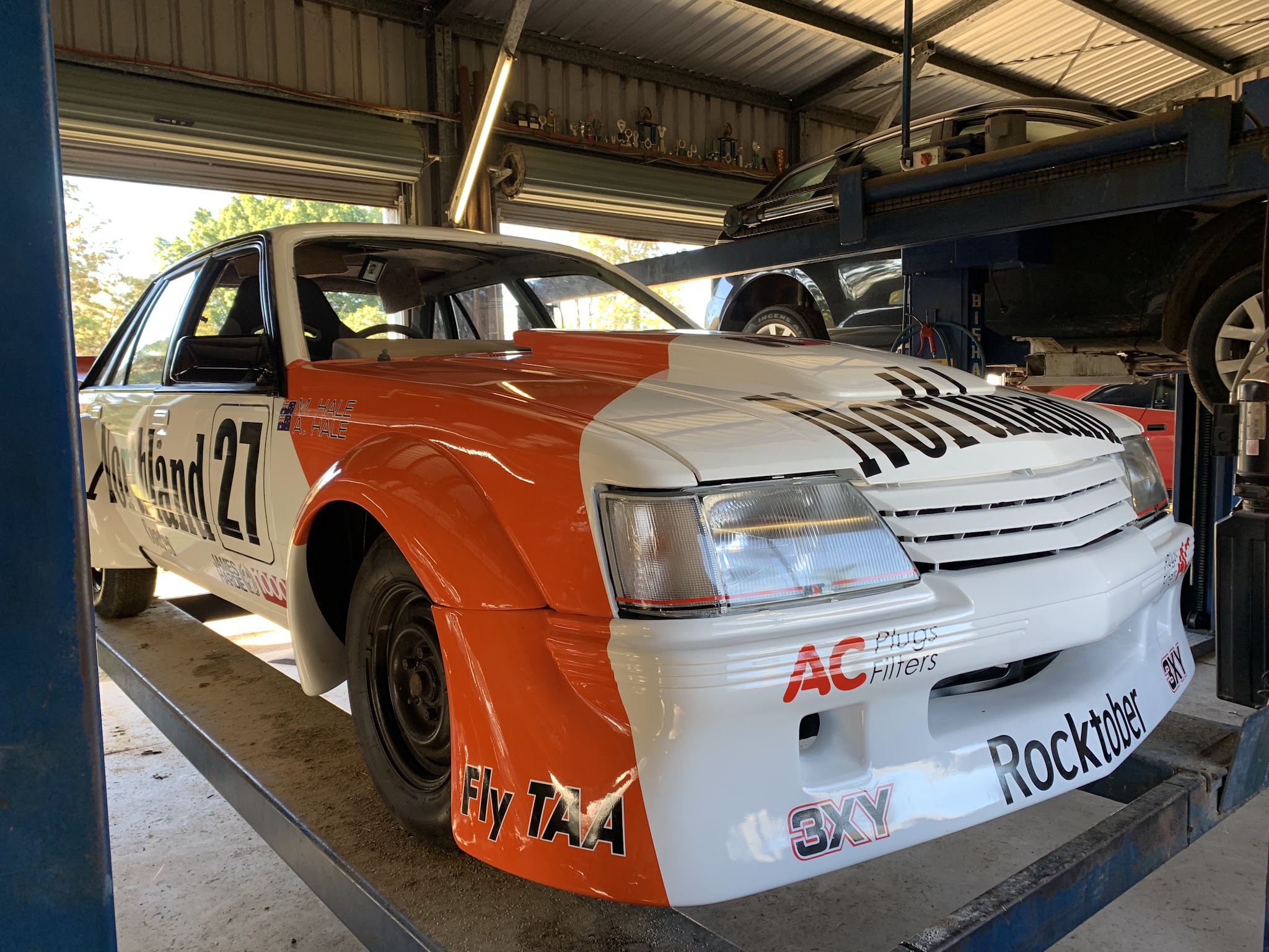 Orange and white Group A liveried VK Commodore parked inside the workshop beside the hoist