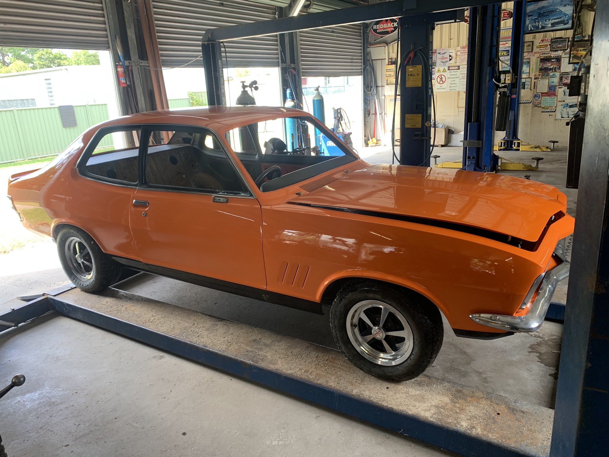 An orange LJ Torana coupe sitting on the ramps of the two post hoist inside the workshop, roller doors open behind it