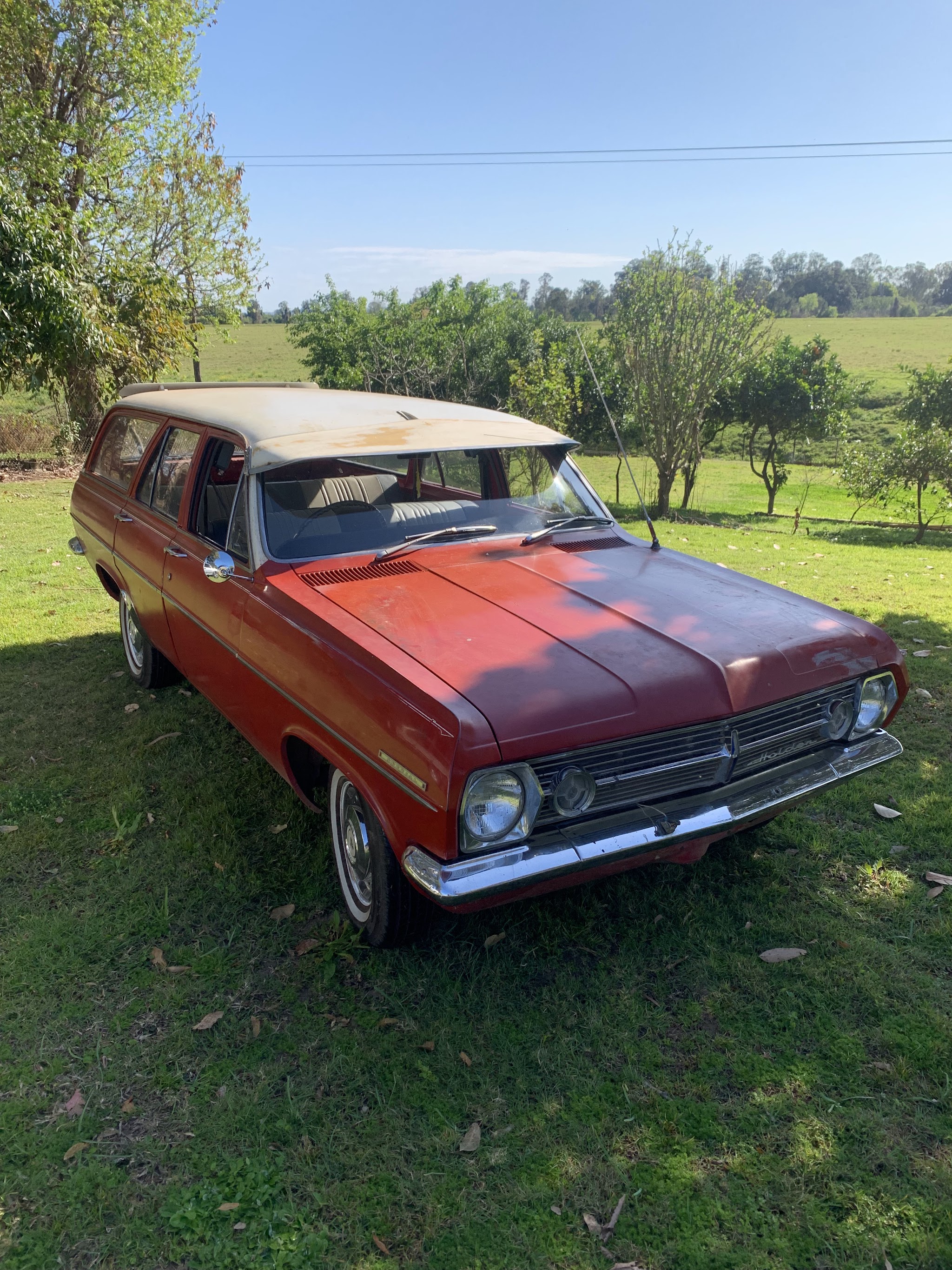 A two tone red and cream Holden HR station wagon parked on a lawn with paddocks behind it
